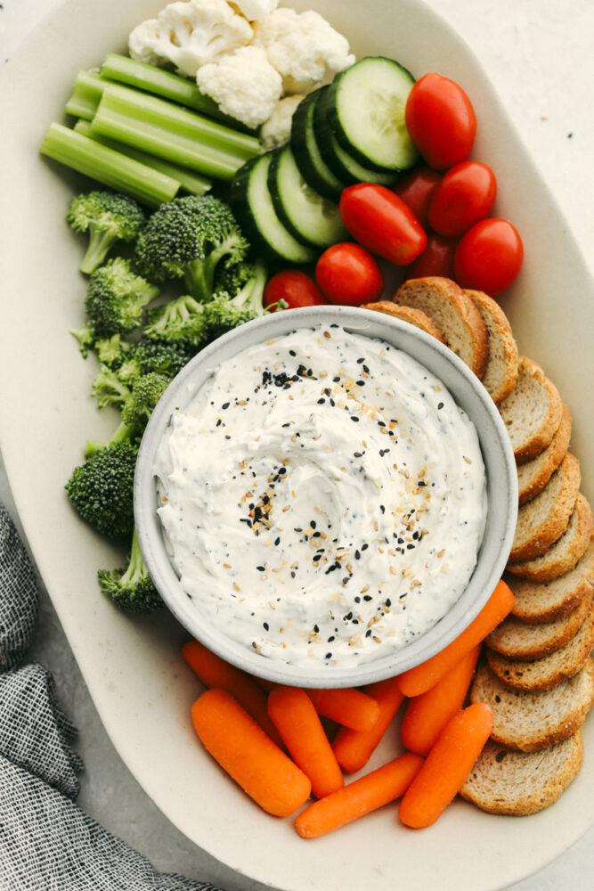 A platter with veggies and bread and a bowl of everything bagel dip. 