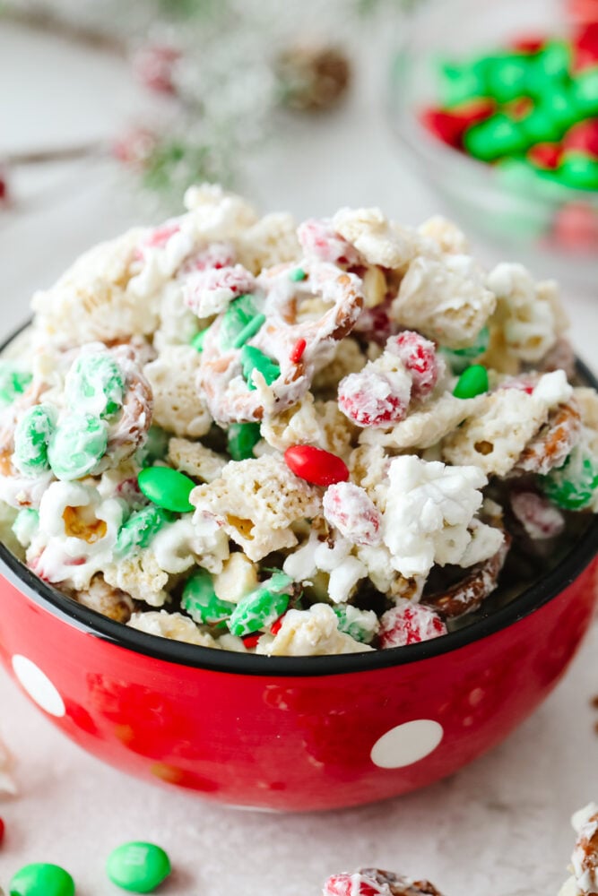Chex, pretzels, m&m's, peanuts, and popcorn covered with white chocolate and sprinkles in a red, polka dot bowl. 