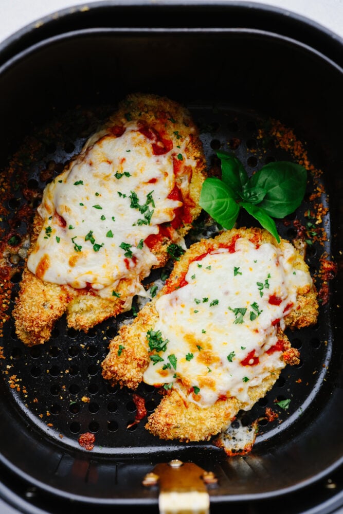 2 pieces of chicken parmesan topped with herb garnish in an air fryer basket.