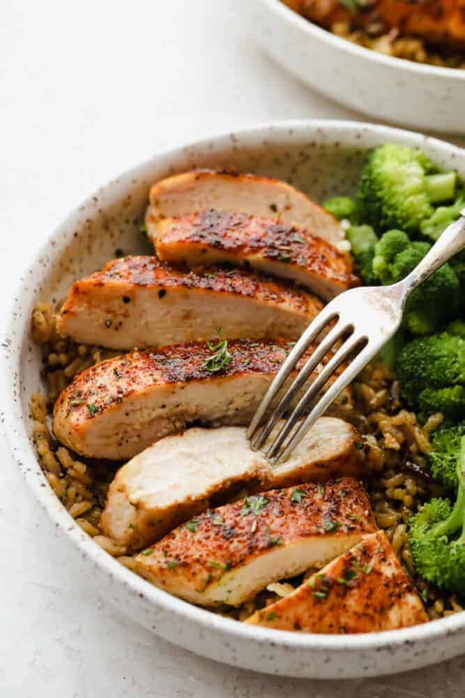 A bowl of cooked chicken and broccoli with a fork sticking into a piece of chicken. 