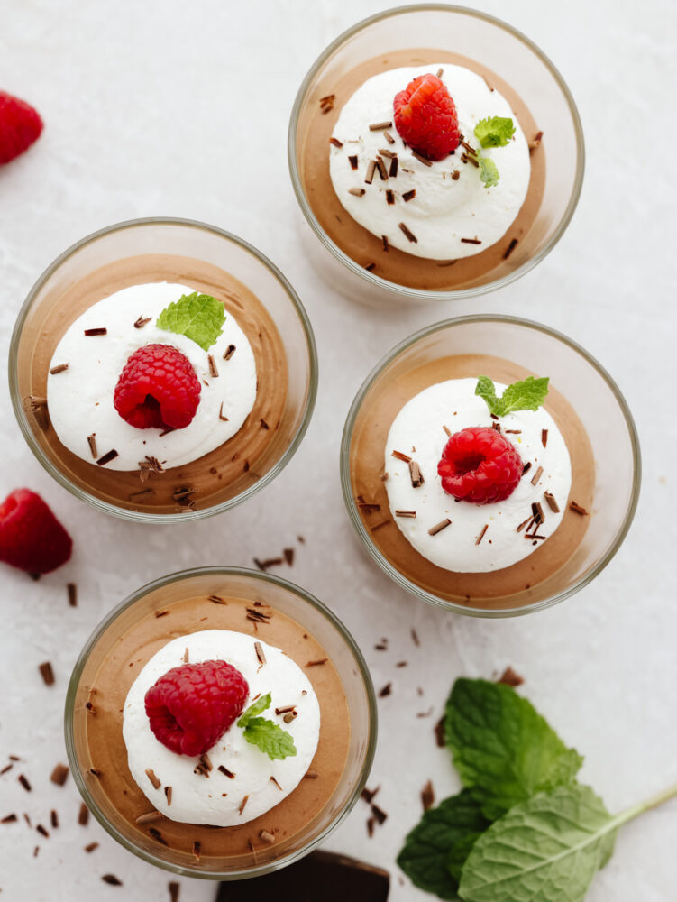 The top view of 3 glasses of chocolate mousse. Each topped with whipped cream, a raspberry and a mint leaf. 