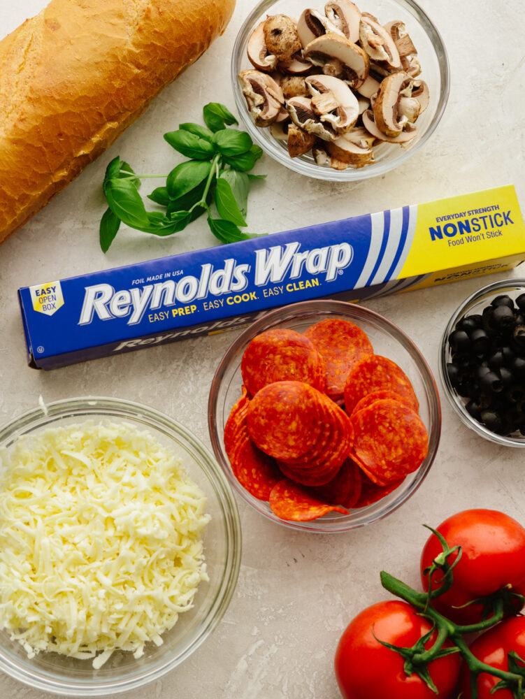 Reynolds Wrap® Non-Stick foil, pepperonis, mushrooms, tomatoes, basil, olives, and cheese in bowls on the counter. 