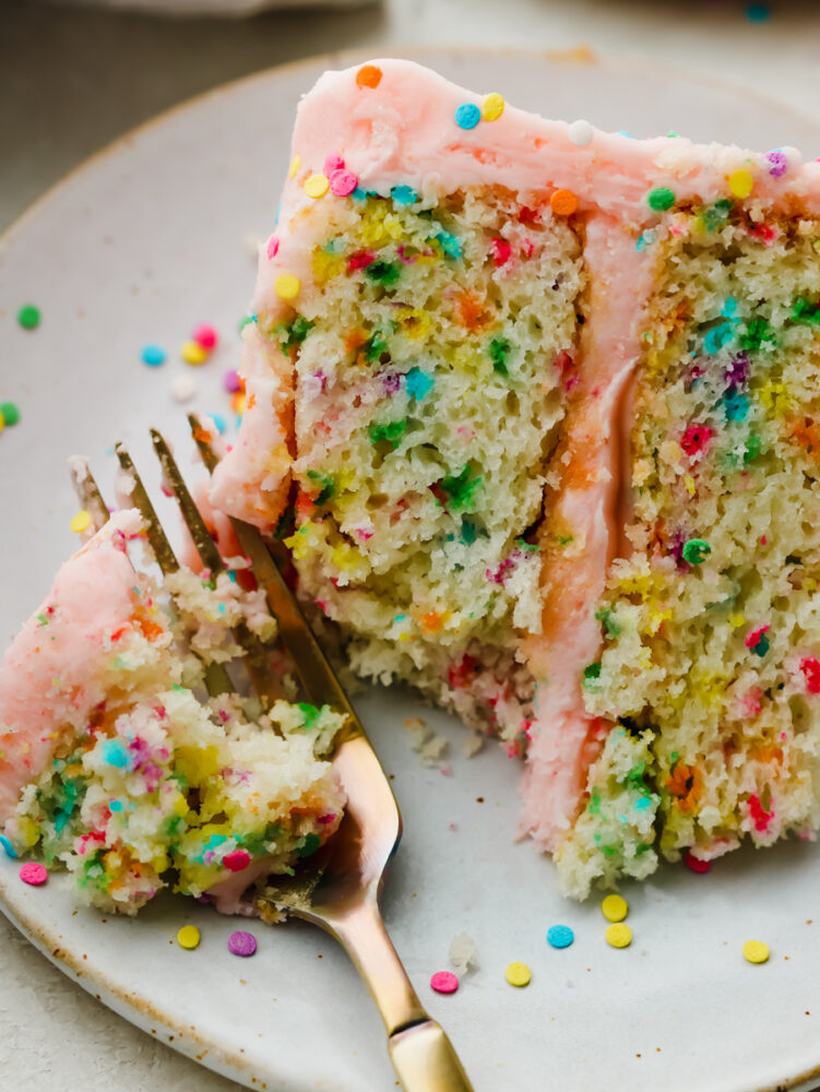 A slice of funfetti cake on a plate being cut with a gold fork. 