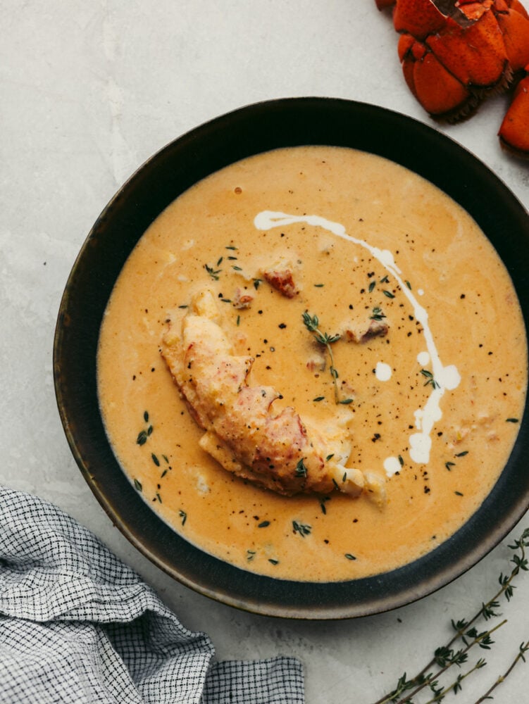 A bowl of lobster bisque with garnish and a lobster tail in it. 