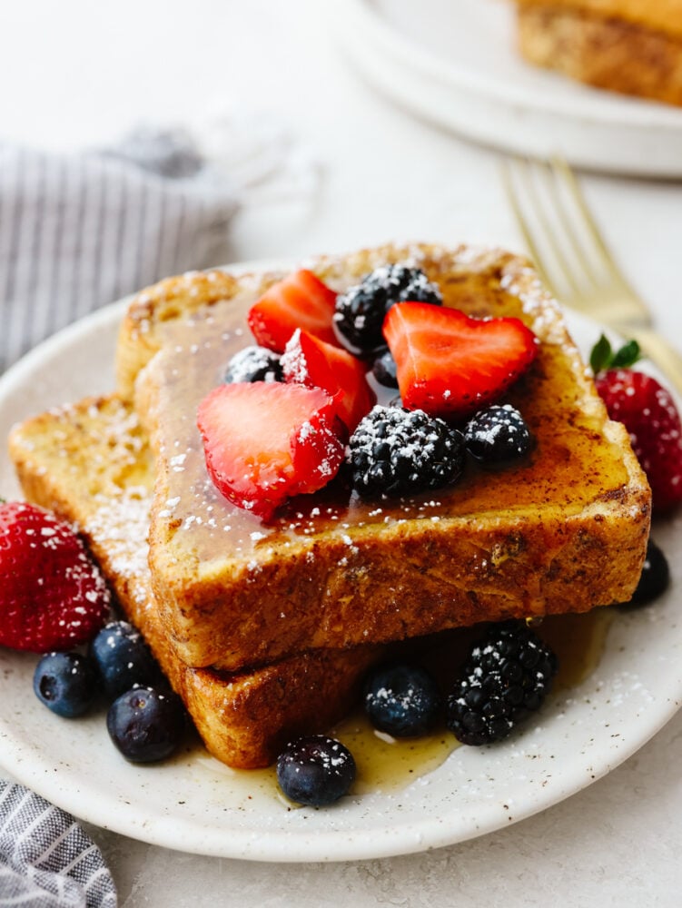 A plate with a stack of french toast that is topped with syrup, berries and powdered sugar. 