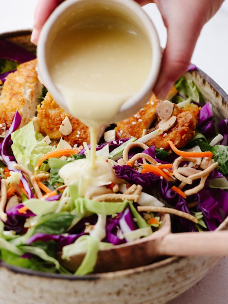 A bowl of asian chicken salad with a creamy vinaigrette being poured on top. 