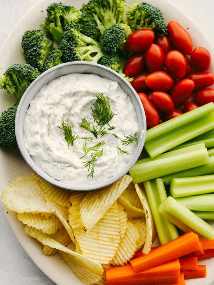 A plater with dill dip in a bowl surrounded by chips, celery, tomatoes and broccoli. 