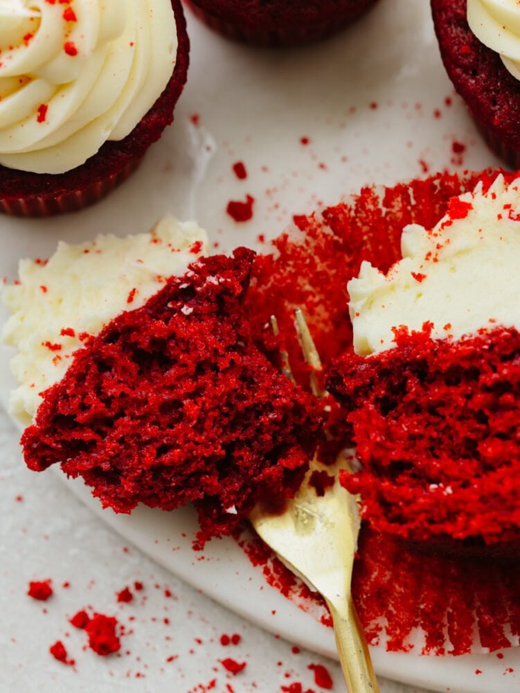 A red velvet cupcake being cut into with a gold fork on a white plate. 