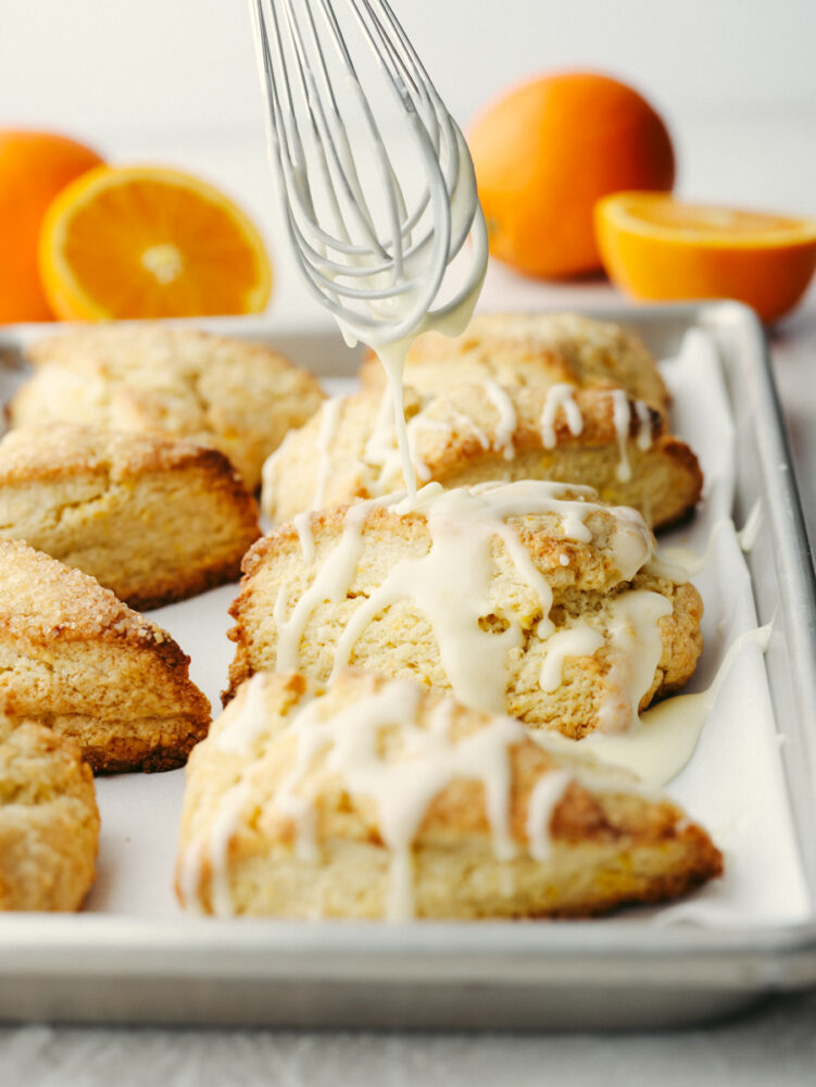 Orange glaze being drizzled with a whisk onto an orange scone. 