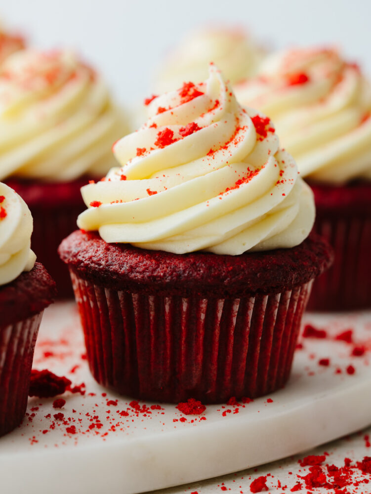 A red velvet cupcake on a platter with cream cheese frosting and red velvet crumbs on top. 