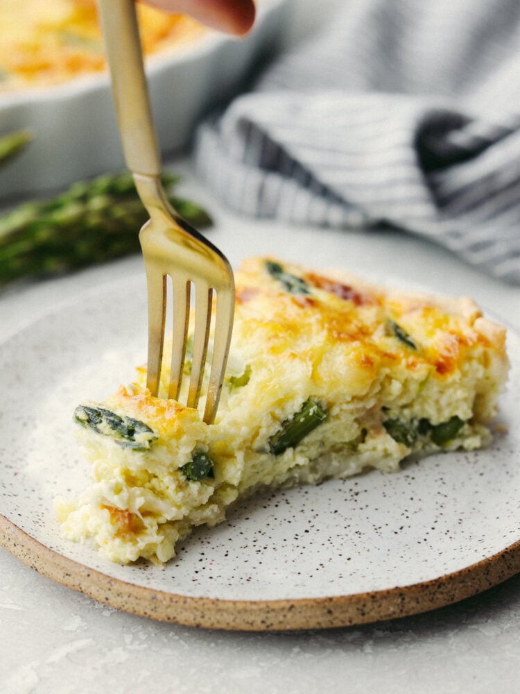 A slice of asparagus quiche on a plate with a gold fork cutting into it. 