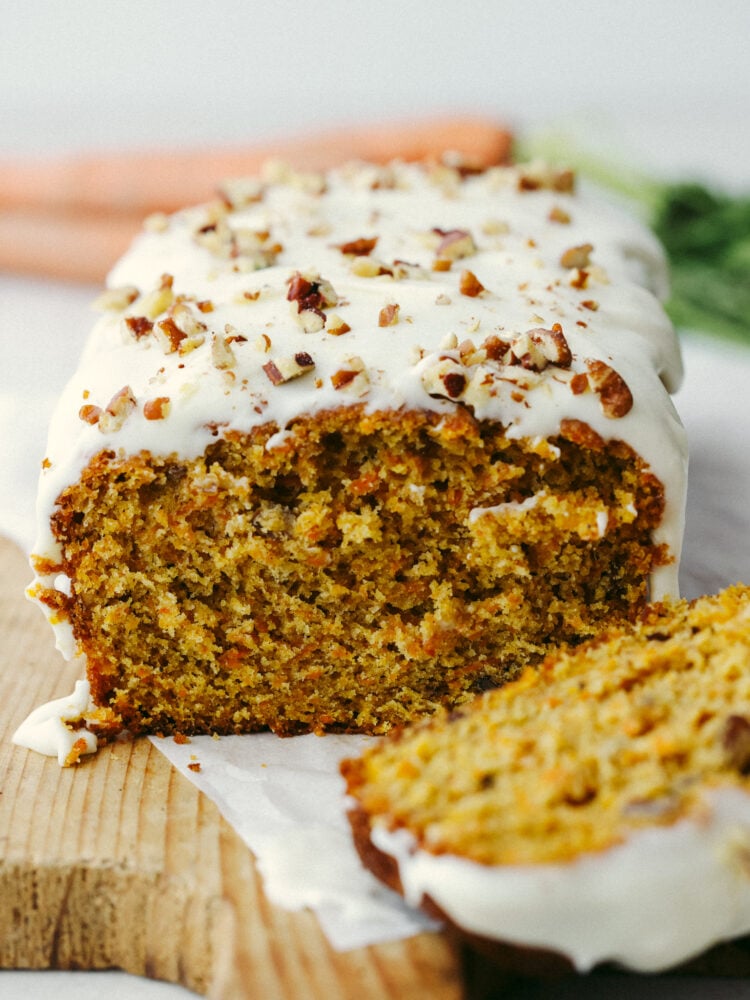 A loaf of carrot cake bread with a slice taken out and a glaxe and pecans on top. 