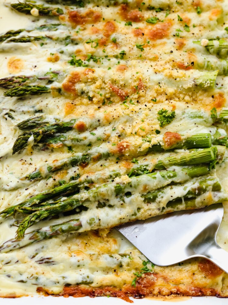 A close up of baked asparagus with cheese and a serving spoon. 