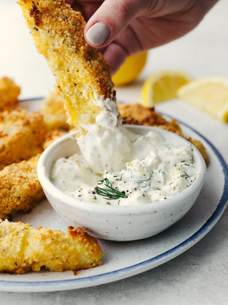 A fish stick being dipped into a small white bowl filled with tartar sauce. 