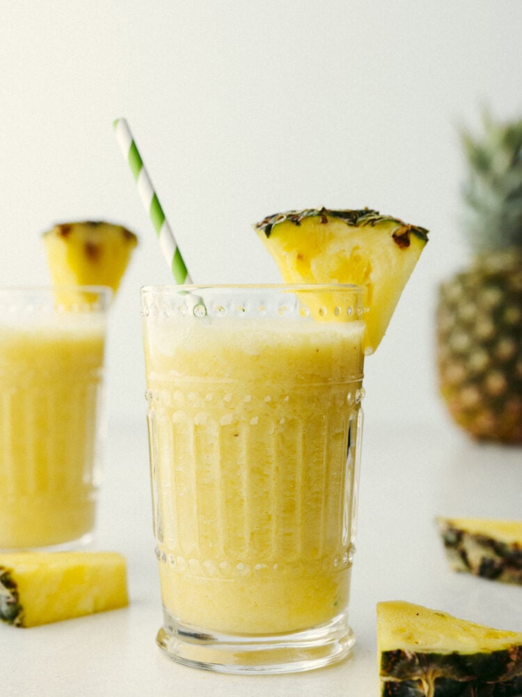 A pineapple smoothie in a glass with a green and white straw and a slice of pineapple on the glass. 