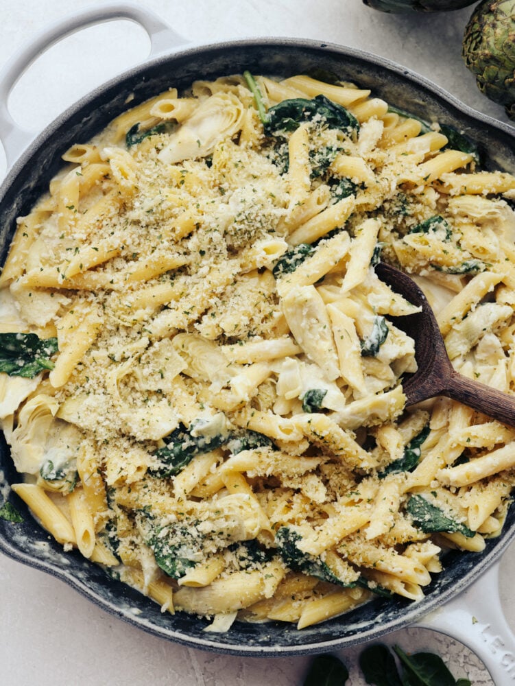 A skillet filled with spinach artichoke pasta and topped with some bread crumbs.