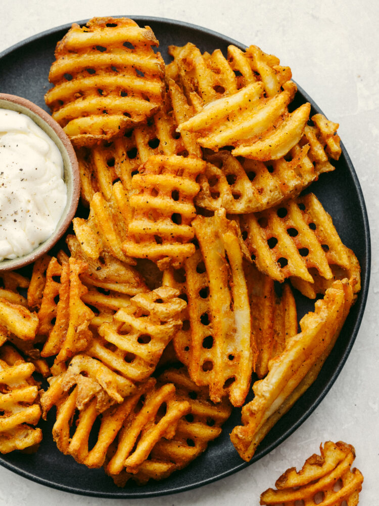 Waffle fries on a black plate with a small bowl of garlic aioli sauce. 