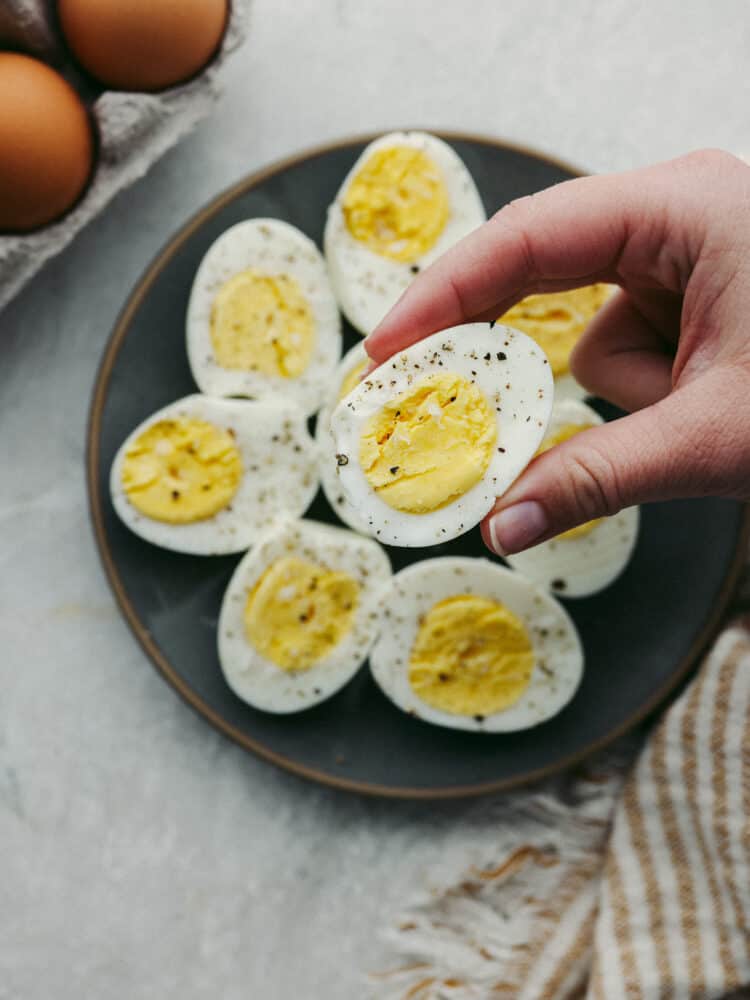 Someone holding the half of a sliced hard boiled egg above a plate filled with hard boiled eggs, sliced and ready to eat. 