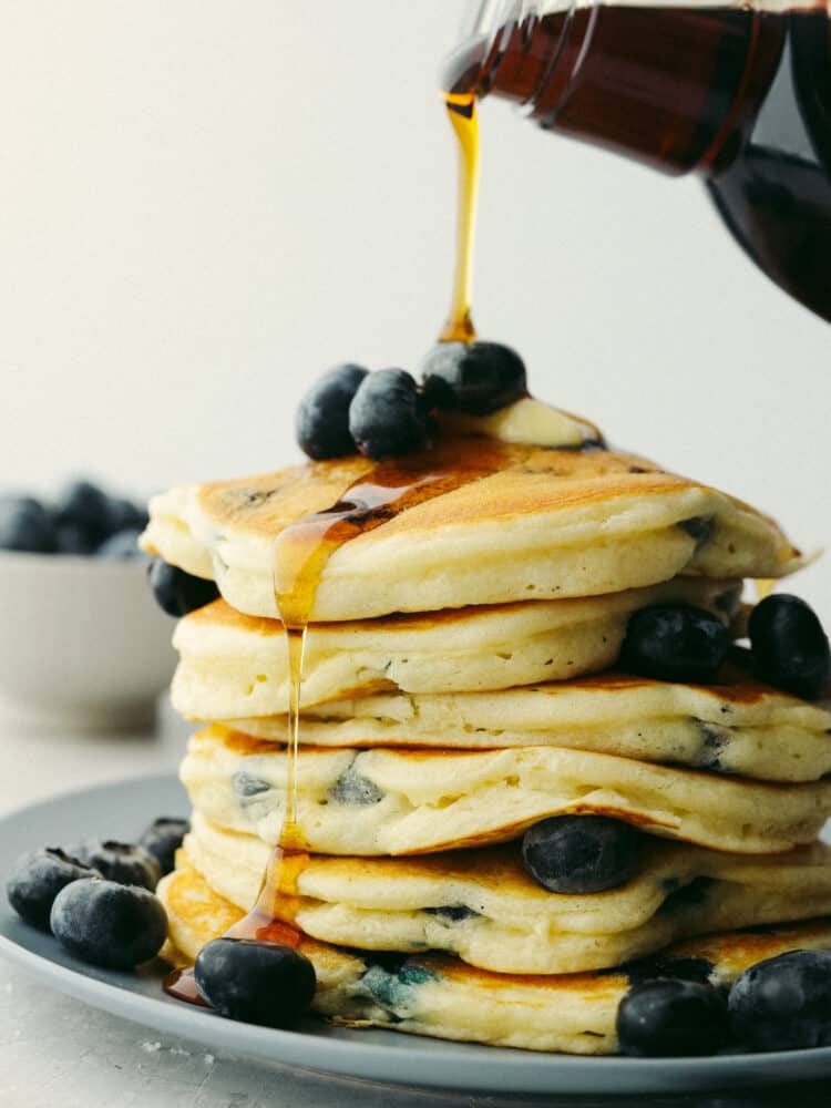 Syrup being poured onto a stack of blueberry pancakes. 