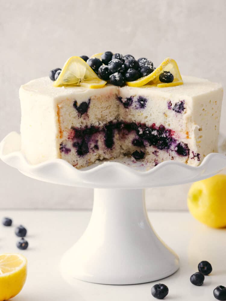 A lemon blueberry cake on a white platter with a quarter of it cut out so that you can see the inside. 
