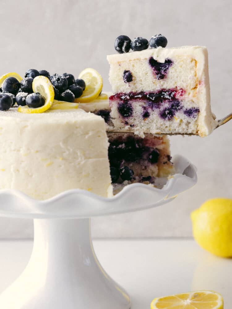 The side view of the lemon blueberry cake on a platter with a slice being taken out. 