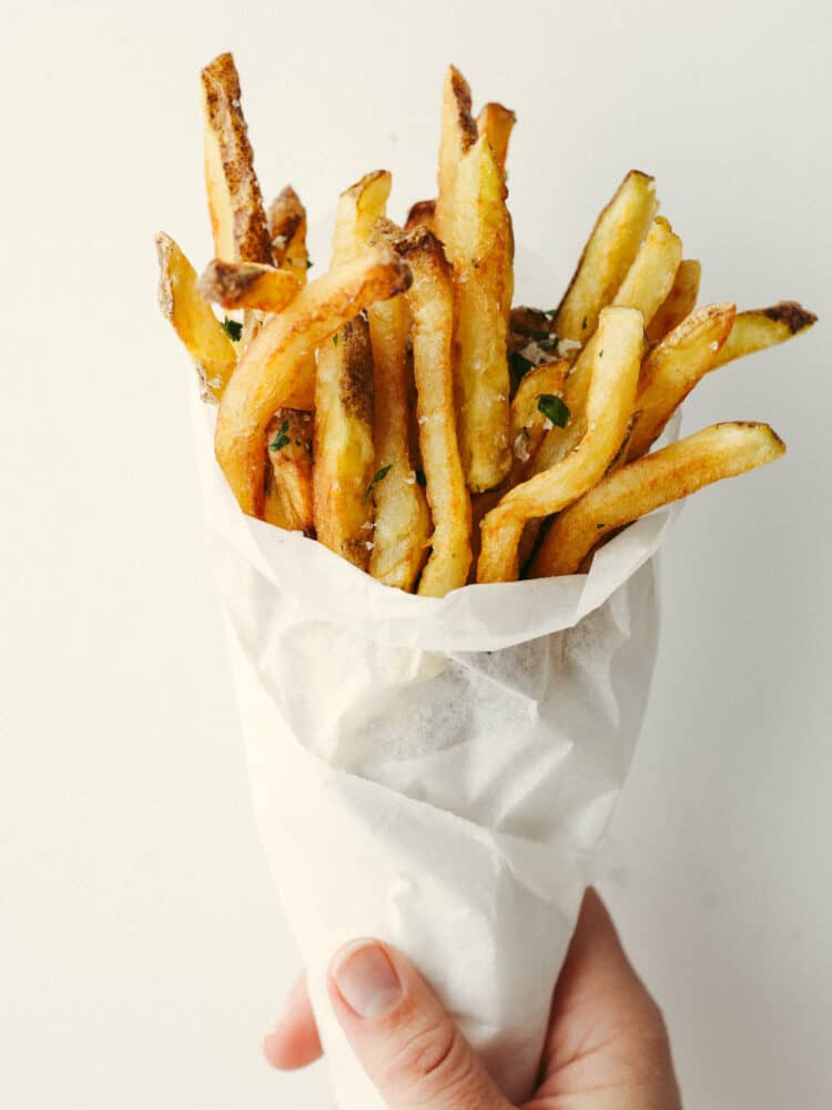 Cooked Pommes Frites in a cone of parchment paper being held by a hand. 