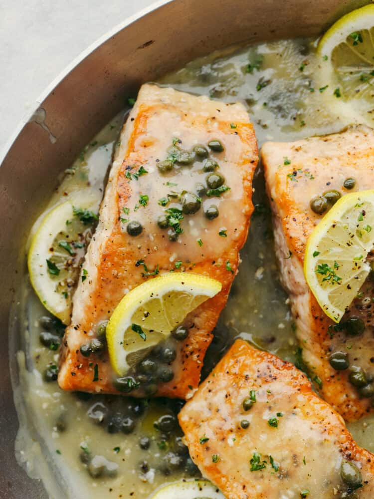 A close up salmon piccata in a pan garnished with capers and lemon slices. 