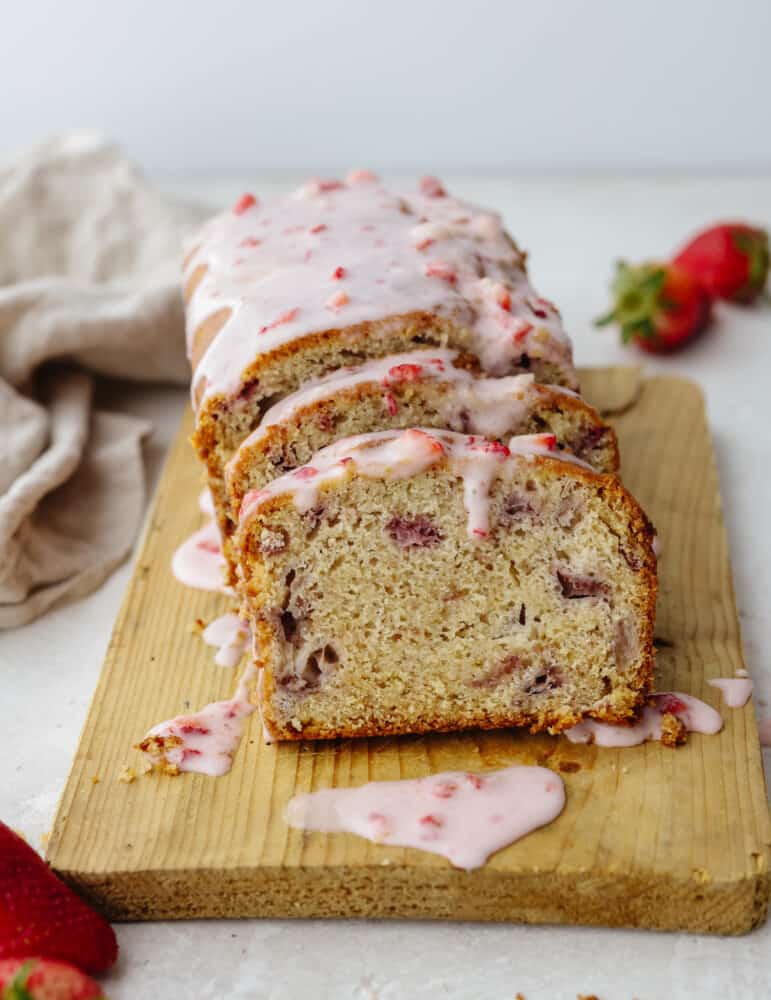 A loaf of strawberry quick bread sliced with a strawberry glaze sitting on a wooden cutting board. 