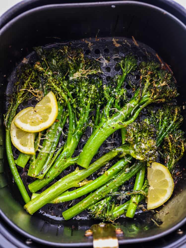 An air fryer basket with cooked broccolini and some lemon slices. 