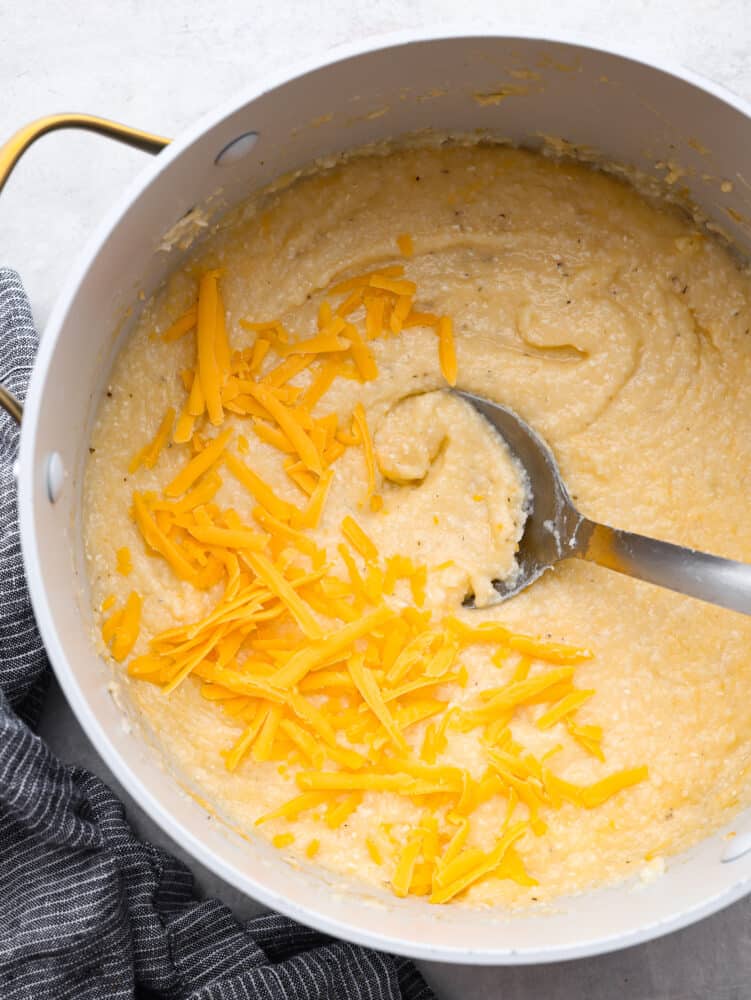 Top view of a large pot of cheesy grits with a large metal spoon laying in the grits. Shredded cheddar cheese garnished on top with a gray kitchen bowl on the side of the pot.