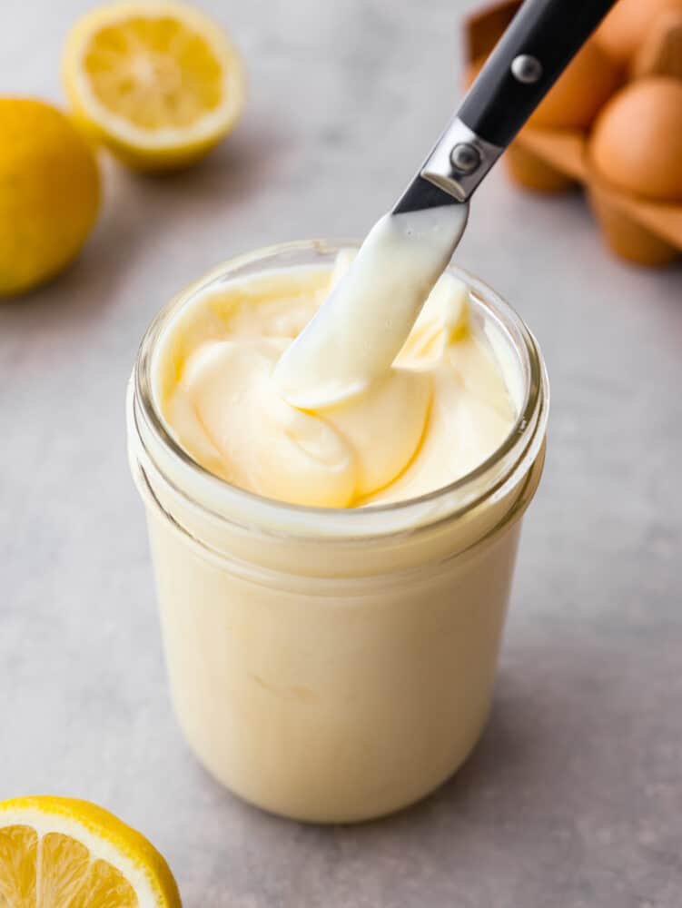 Close-up foto van romige mayonaise in een glazen pot met een mes dat mayonaise opheft.  Citroenen en eieren op de achtergrond.