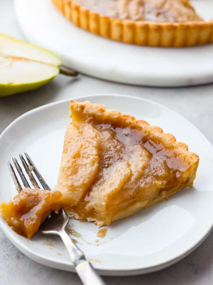 A slice of pear tart on a plate with a silver fork cutting a piece of it. 