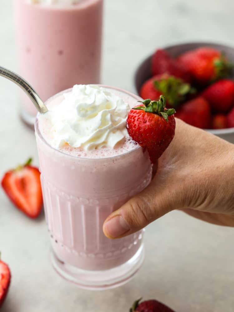 A hand holding a glass filled with strawberry milkshake. There is a silver straw in the cup and a fresh strawberry on the side of the glass. 