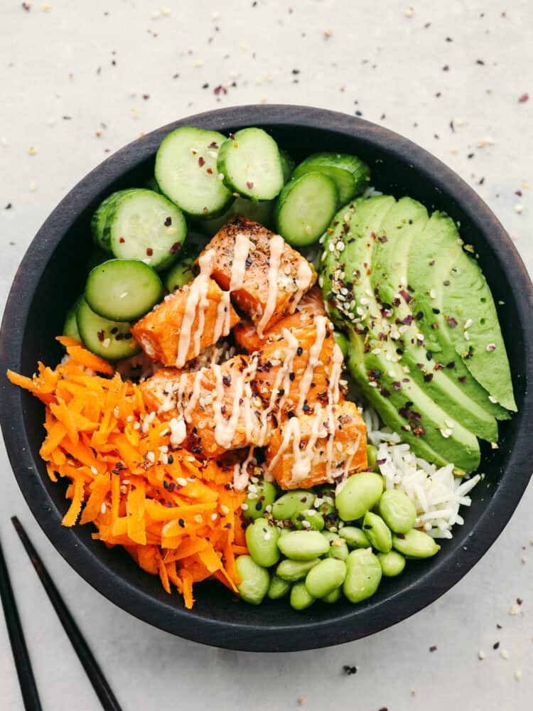 Overhead photo of the salmon rice bowl in a wood bowl.  Ingredients in the bowl is rice, cut up cucumber, sliced avocado, shredded carrots, and edamame.  Sriracha mayonnaise is drizzled on top with a sprinkle of sesame seeds.