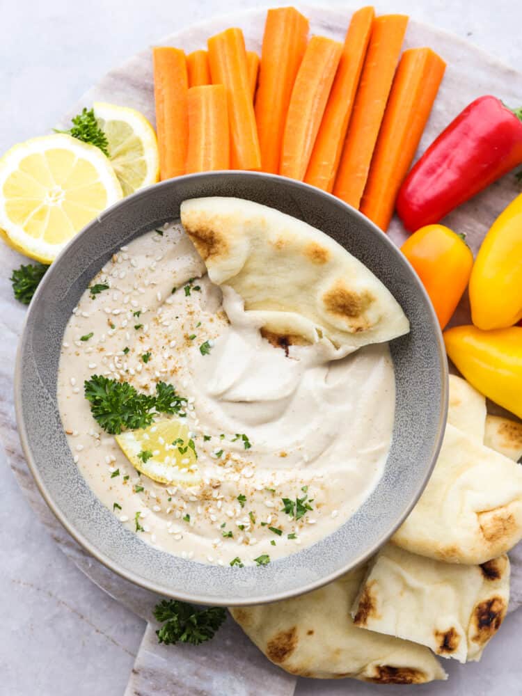 The top view of a bowl with tahini sauce garnished with parsley and sesame seds and a slice of pita bread being dipped into it. 