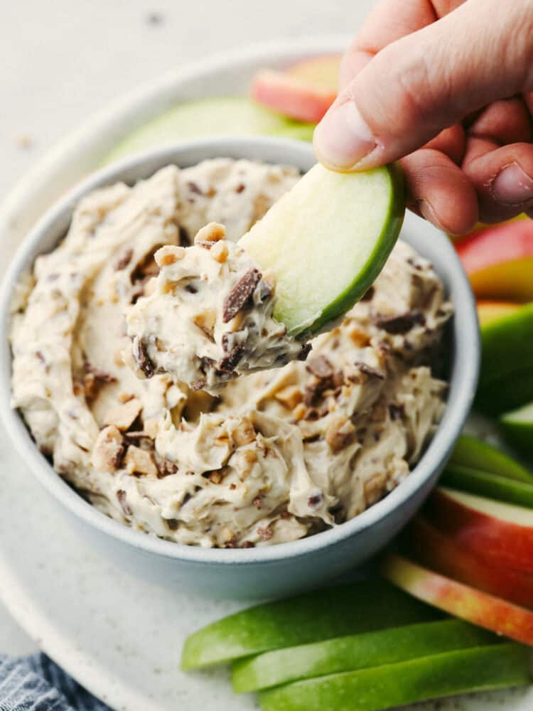 A close up of someone dipping an apple slice into apple toffee dip. 