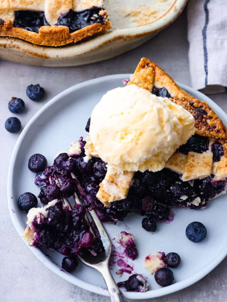 A slice of blueberry pie with a scoop of vanilla ice cream on a periwinkle colored plate.
