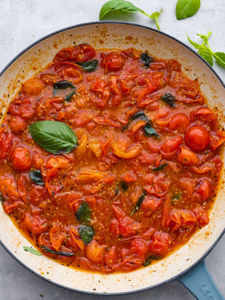 Overhead photo of cherry tomato sauce in a blue skillet with fresh basil on the side.
