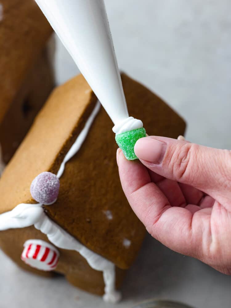 A hand piping royal icing onto the bottom of gumdrops with a gingerbread house in the background.