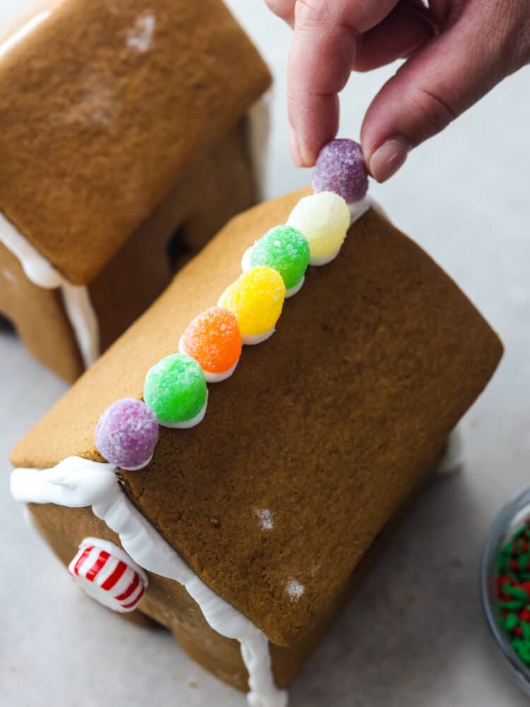 A hand placing the gum drops on top of the roof of a gingerbread house.