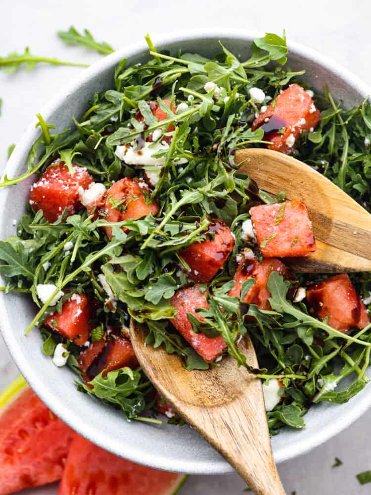 Top view of watermelon arugula salad in a large gray bowl.  Wood serving spoons are sitting in the bowl of salad with watermelon and arugula scattered on the counter next to the bowl.