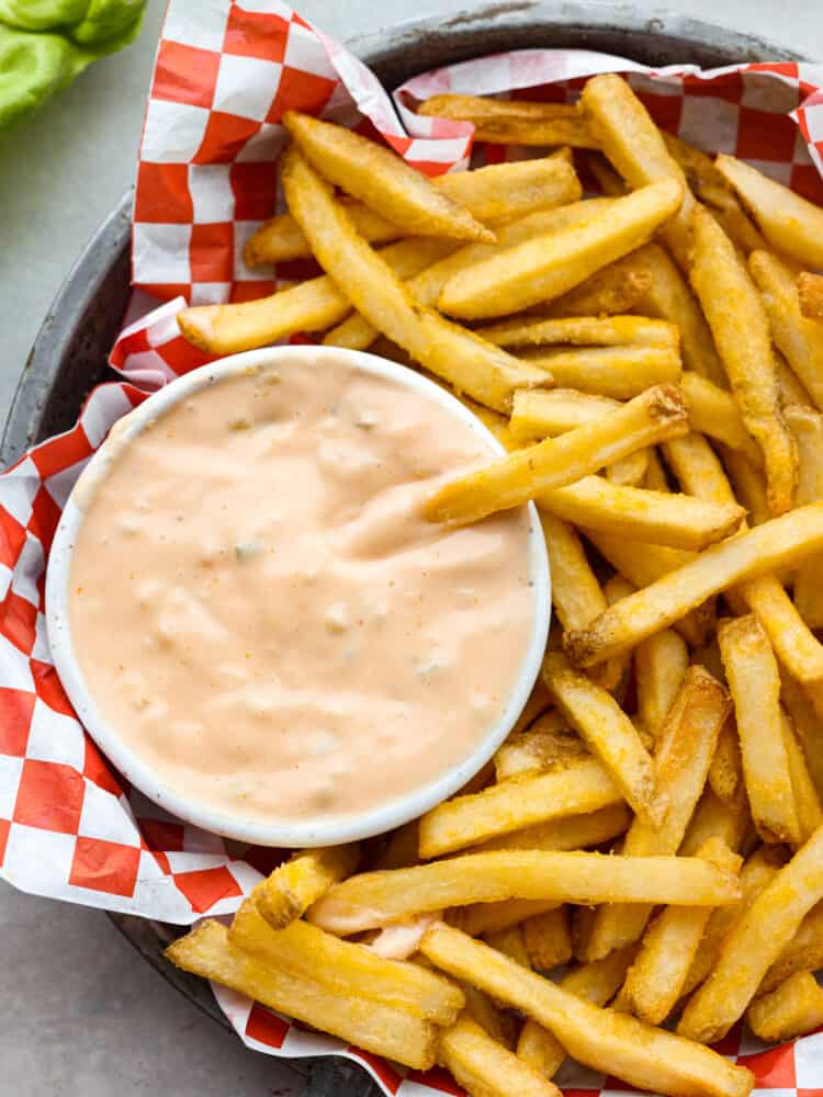 Overhead photo of a French fry dipping in a bowl of sauce. The small bowl of sauce and extra French fries are inside of a metal tin pan. The metal tin pan is lined with red and white checkered parchment paper.