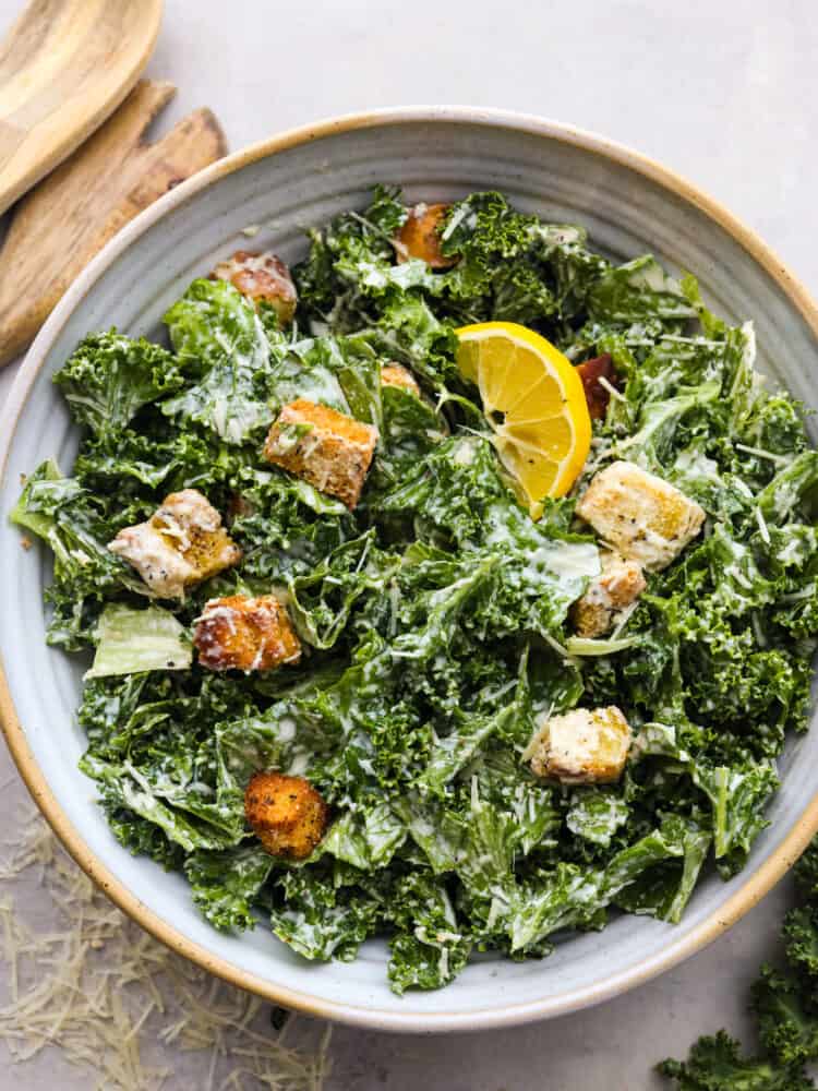 Top view of kale Caesar salad in a large bowl garnished with a lemon wedge.  Wood serving spoons on the side.