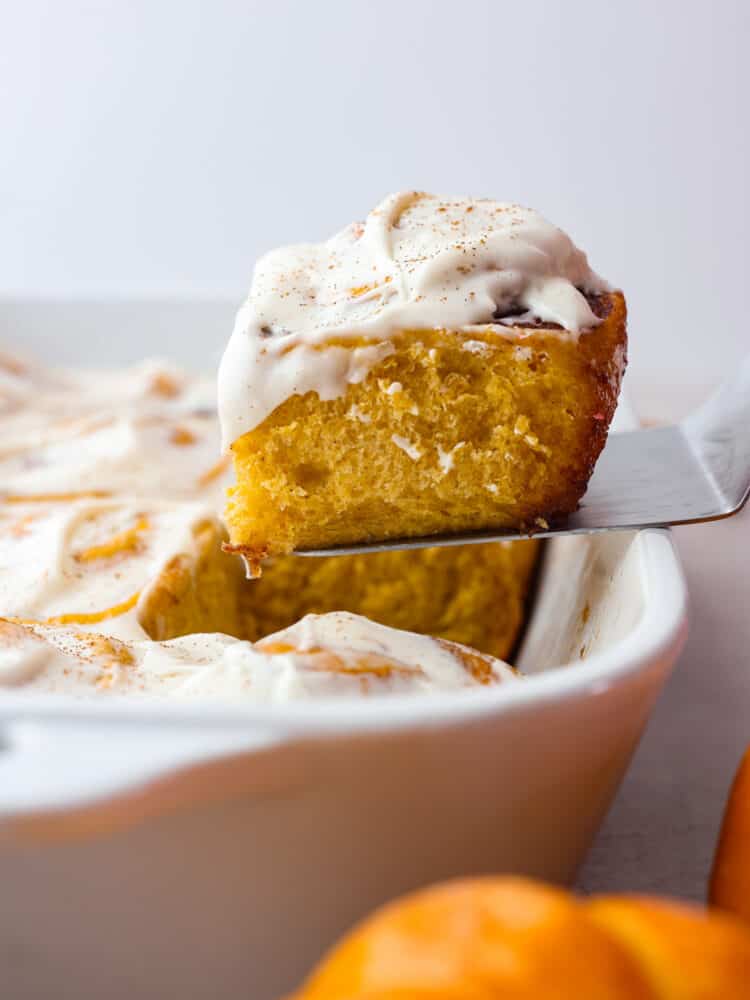 A cinnamon roll being taken out of a white pan with a metal spatula.