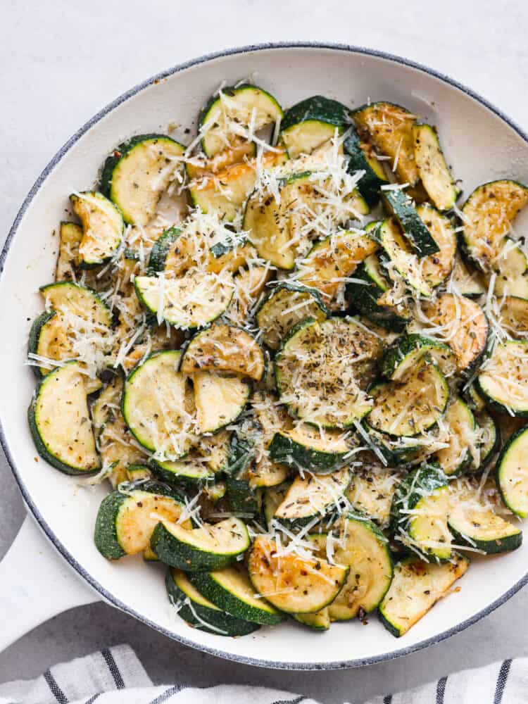 Top view of sautéed zucchini and melted parmesan cheese on top.  The sautéed zucchini is beautifully displayed in a large white skillet with a striped towel next to it.