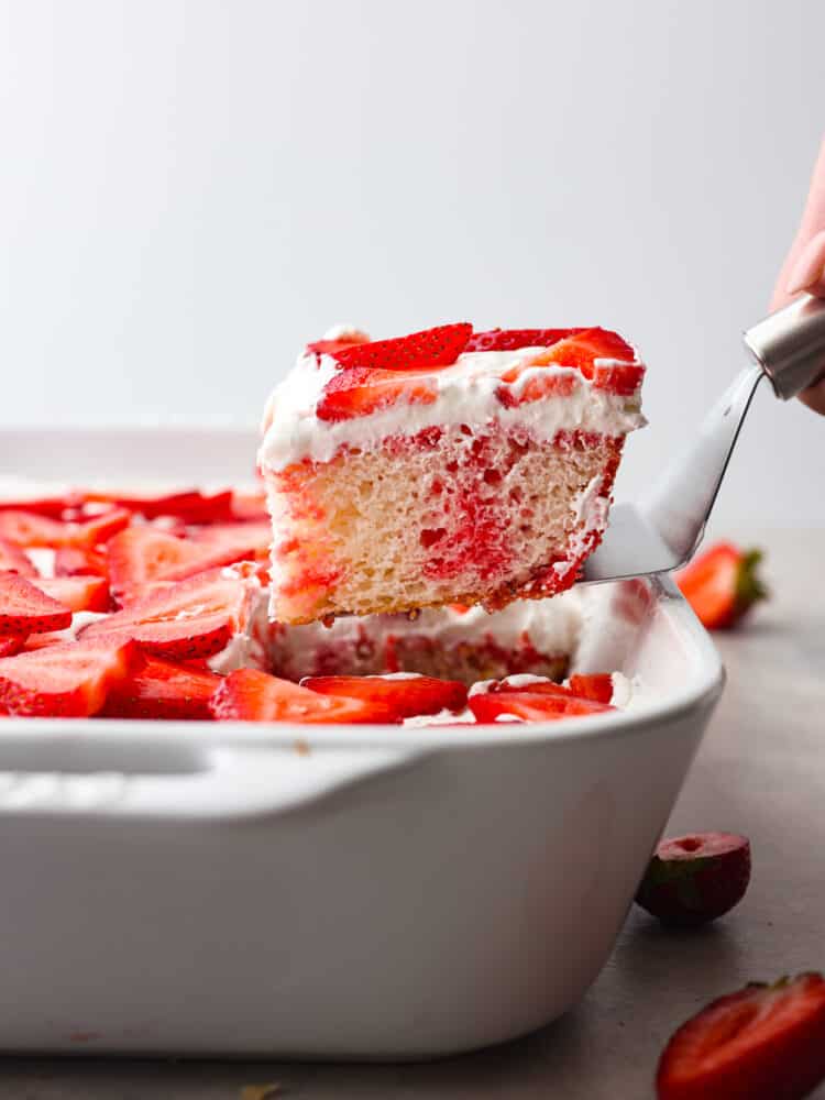 The side view of a slice of strawberry poke cake being taken out of the pan with a silver cake server. 