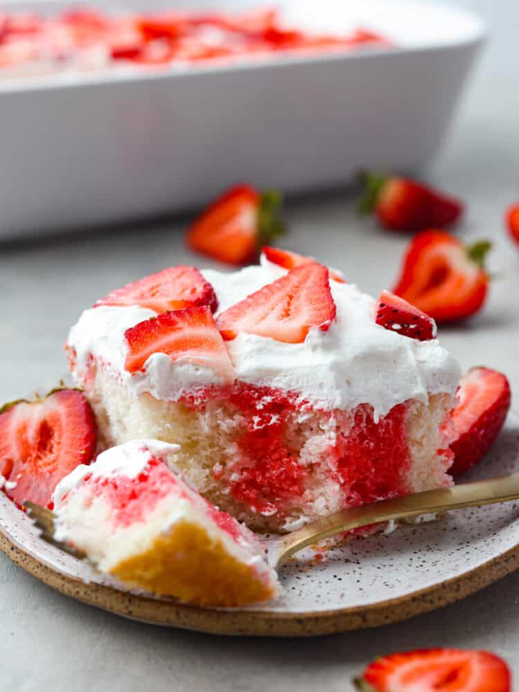 A piece of strawberry poke cake on a plate with a gold fork cutting a bite. 
