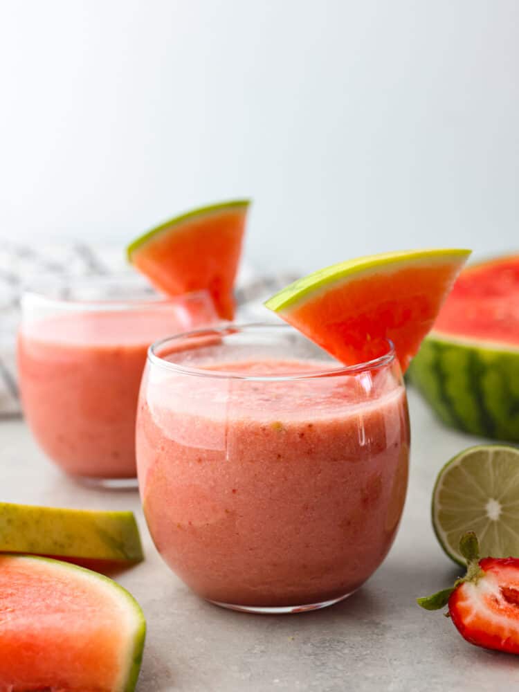 A glass cup filled with watermelon smoothie and a watermelon slice on the side of the cup. 