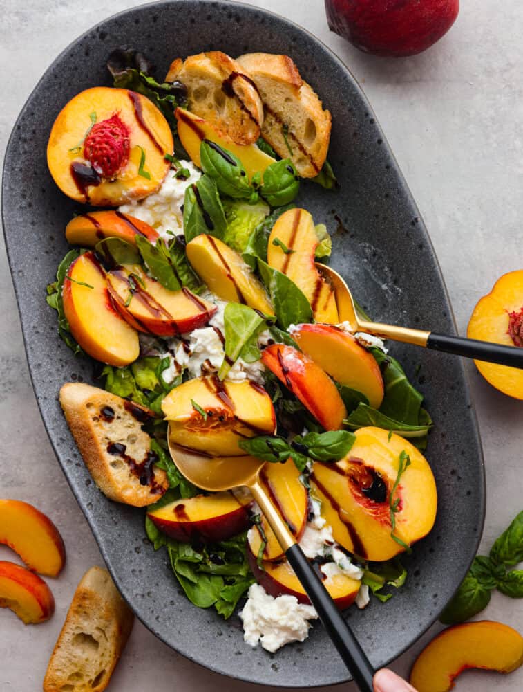 Top view photo of peach burrata salad on a gray platter.  Black and gold serving spoons are lifting up the salad. Sliced peaches and crostini garnished on the side.