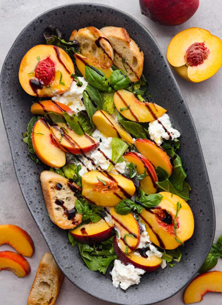 Top view photo of peach burrata salad on a gray platter.  Balsamic glaze and basil garnished on top. Sliced peaches and crostini garnished on the side.  One whole and halved peach next to the platter.